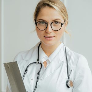 Smiling female healthcare professional in white coat holding clipboard indoors.